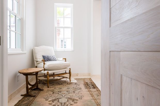 A bright, narrow sitting nook viewed through a partially open whitewashed timber door. The room has white walls, tall multi-pane windows, pale timber flooring, a vintage upholstered armchair with wooden arms, a small round timber side table, and a muted patterned area rug. The space feels quiet, airy, and traditionally styled.