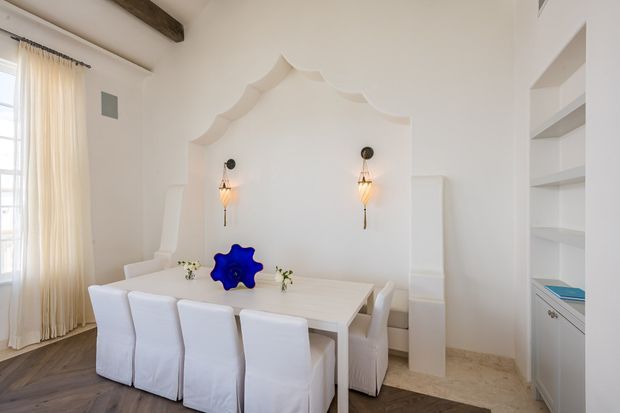 Bright, refined dining room with a long white dining table and upholstered slipcovered chairs, set against a sculptural white plaster feature wall with an ornate scalloped arch and two decorative wall sconces. Built-in open shelving and cabinetry occupy the right side, while tall sheer curtains admit abundant daylight on the left. A dark exposed timber ceiling beam and dark timber flooring add contrast to the predominantly white interior.