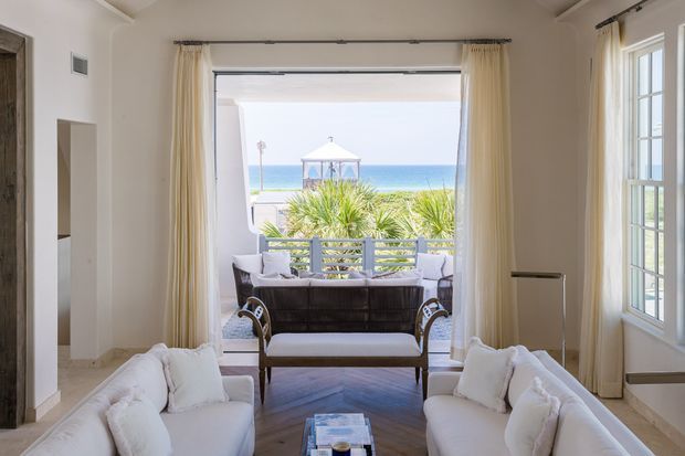 Bright coastal living room viewed toward an open balcony or terrace, with two white sofas, a dark leather sofa, a central upholstered coffee table, pale curtains, and herringbone timber flooring. Large open glazing frames a landscaped seaside view with a pavilion and ocean beyond. The room has a refined, airy, symmetrical arrangement and tall ceilings.