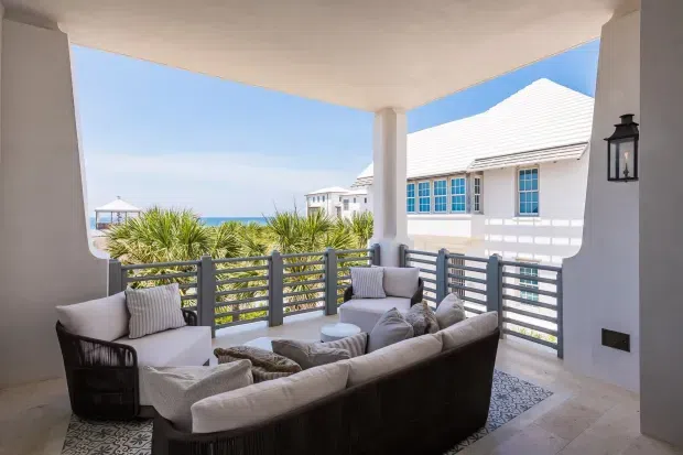 A covered coastal outdoor lounge terrace with a large upholstered sectional sofa, armchairs, and a low coffee table arranged around a patterned rug. White rendered columns and ceiling frame the space, with grey horizontal metal railings, pale stone-look flooring, and a lantern wall light. Tropical landscaping, blue sky, ocean, and a neighbouring white-roofed building are visible beyond the terrace.