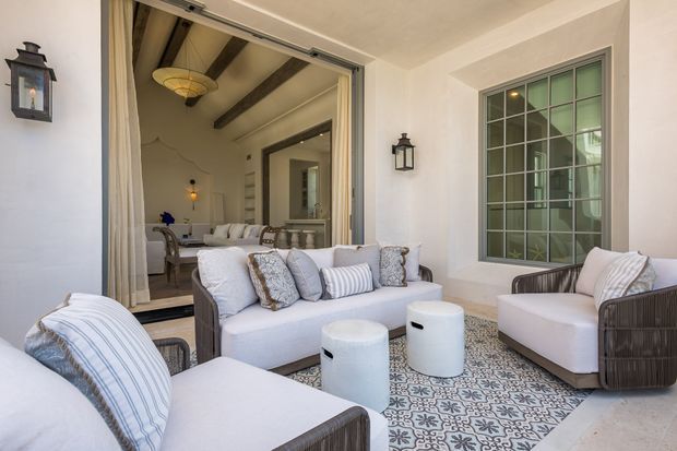 Covered outdoor sitting area furnished with a white upholstered sofa, armchairs, ottomans, and patterned rug. Large bi-fold or sliding glazed doors open directly into an adjoining interior living space, while a tall gridded window, wall-mounted lanterns, exposed dark ceiling beams, and pale rendered walls define the architecture. The palette is light and warm with Mediterranean-inspired detailing and a relaxed, refined atmosphere.