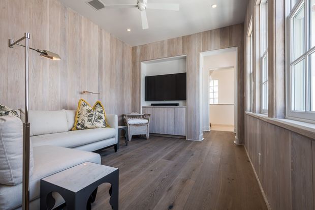 A compact, refined living room with full-height light timber wall panelling, matching timber flooring and built-in media cabinetry. A pale upholstered sofa, patterned accent cushions, sculptural dark side table, and contemporary floor lamp sit beside large multi-pane windows that bring in abundant natural light. A doorway leads to an adjacent hall or room.