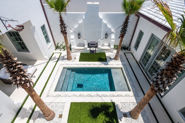 A bright, symmetrical courtyard viewed from above, centered on a compact rectangular swimming pool. White rendered walls with tall glazed doors and windows enclose the space, while palm trees, artificial-looking grass panels, white gravel, and decorative patterned paving create a resort-like outdoor setting. A covered seating or entertaining area is visible at the far end.
