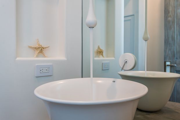 Close-up interior view of a refined bathroom featuring a freestanding white oval bathtub in the foreground, a second freestanding tub or basin behind it, a large mirror, white painted wall panelling, electrical outlets, tall sculptural white decorative fixtures, and a small starfish ornament. The palette is soft white and pale grey with a calm, coastal-inspired feel.
