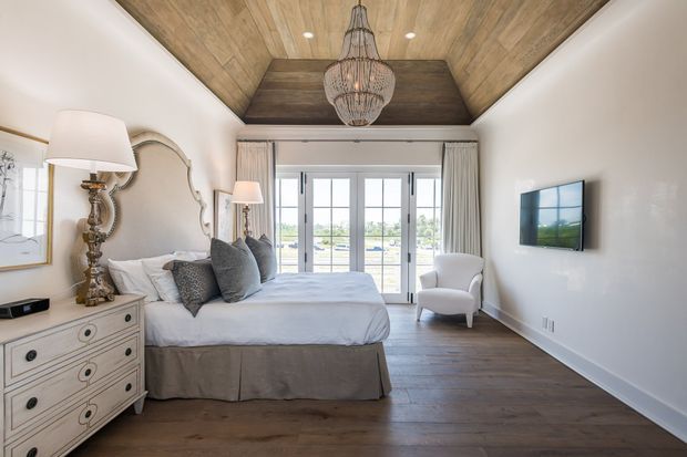 Bright, elegant bedroom with a large upholstered bed, layered neutral bedding, ornate bedside lamp, decorative dresser, armchair, and a wall-mounted television. The room features dark timber flooring, white walls, full-height glazed French doors with curtains, and a striking recessed timber ceiling panel with a woven pendant light. A balcony or exterior view is visible beyond the doors.