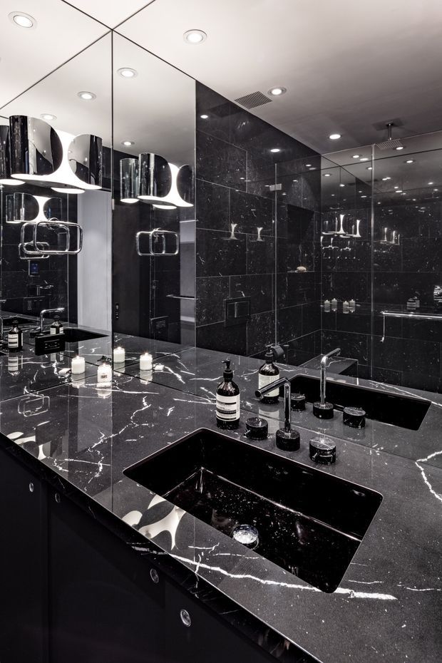 A dramatic, luxurious bathroom vanity viewed close-up. The room features a large black stone vanity benchtop with prominent white veining, two integrated rectangular black basins, chrome tapware, dark cabinetry, black marble-look wall tiles, and extensive mirrored surfaces. Sculptural wall lights and recessed ceiling lights add a polished, high-contrast atmosphere.