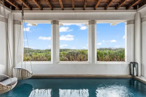 A covered indoor or semi-enclosed swimming pool with deep blue water, white rendered walls, and three large open window bays overlooking tall vegetation. The space features a rustic exposed timber-beam ceiling, structural concrete or rendered columns, and suspended woven lounge chairs at the edge of the pool. Bright daylight and the water create a relaxed resort-like atmosphere.