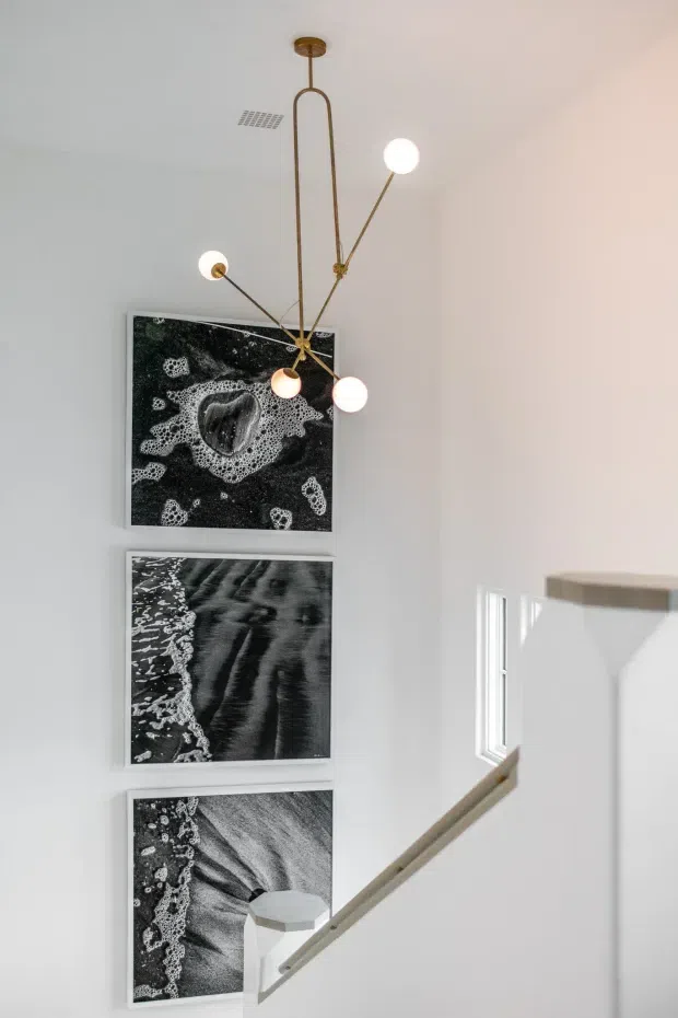 A bright, minimalist stairway or landing with white walls and a white balustrade. Three vertically stacked black-and-white photographic artworks decorate the wall, while a sculptural brass-toned multi-arm pendant with spherical white globes hangs overhead. A small window brings in natural light, creating a clean, refined interior.