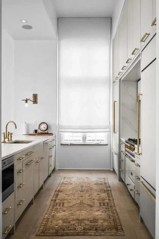A narrow galley kitchen with tall white cabinetry on both sides, integrated appliances, brass hardware and tapware, a marble-look splashback, and light stone or tile flooring. A central window has a white Roman blind, while a vintage runner adds warmth to the otherwise bright, refined space.