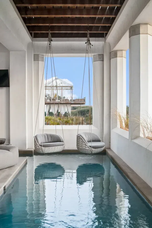 A bright, narrow indoor pool space framed by white plastered columns and large open window bays. Two hanging woven lounge chairs are suspended above the turquoise water, with a dark timber slatted ceiling overhead. Coastal vegetation and a light-filled outdoor structure are visible beyond the openings, creating a serene resort-like atmosphere.