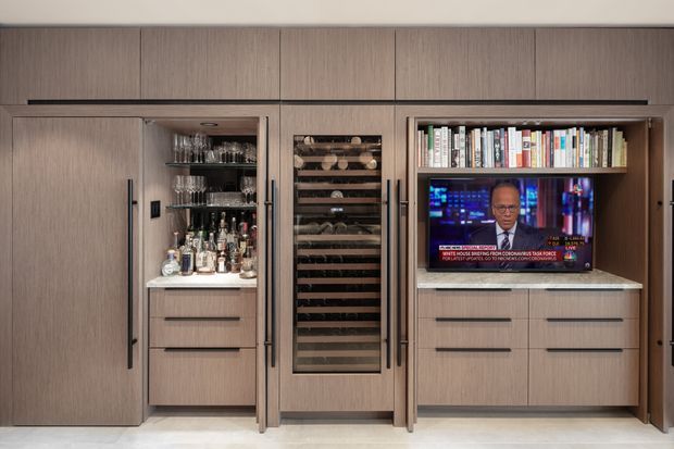 A refined built-in timber-toned wall unit featuring concealed storage, a compact home bar with glassware and liquor bottles, a tall wine rack, and a television set into open shelving beneath a row of books. The cabinetry has clean vertical lines, integrated dark handles, and a warm, contemporary finish.