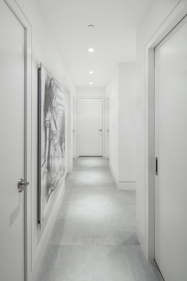 A bright, minimalist white hallway with smooth white walls, multiple flush white doors, recessed ceiling downlights, and a pale grey polished concrete or stone-look floor. A large monochrome framed artwork hangs along the left wall, while a glazed or reflective opening is visible on the right.