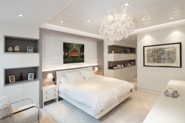 A bright, refined bedroom with a large upholstered bed centered against a textured white feature wall. Built-in white cabinetry and open shelving flank the bed, while bedside lamps, a framed artwork, recessed ceiling lights, and a sculptural crystal chandelier complete the room. A white desk or console occupies the foreground, and the palette is predominantly white, cream, and light grey.