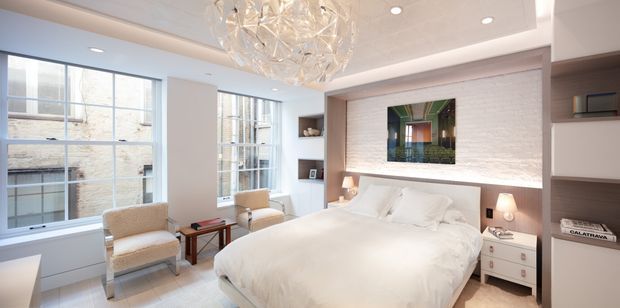 Bright contemporary bedroom with a large upholstered bed, white bedding, two armchairs, expansive multi-pane windows, built-in shelving, and a textured white brick feature wall. A sculptural faceted pendant light provides a decorative focal point, while integrated bedside storage and concealed cabinetry create a refined, clean-lined interior.