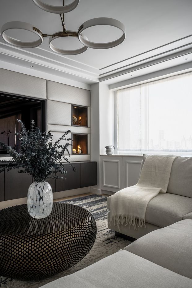 A refined contemporary living room in a monochromatic palette, featuring a large sectional sofa, textured throw, rounded black upholstered coffee table, patterned rug, built-in dark cabinetry with illuminated display niches, a wall-mounted television, decorative wall panelling, layered recessed ceiling details, and a sculptural circular pendant light. Large sheer-covered windows provide abundant filtered daylight.