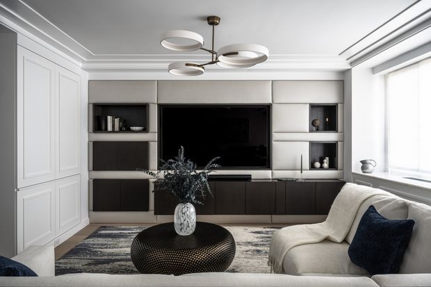 A refined contemporary living room with a built-in wall unit combining a large black television, dark lower cabinetry, pale upholstered or paneled upper sections, and open display niches. A curved cream sofa faces a rounded black coffee table, with a textured neutral rug, navy accent cushion, white panelled storage cabinetry, large windows, and a sculptural multi-arm ceiling light.