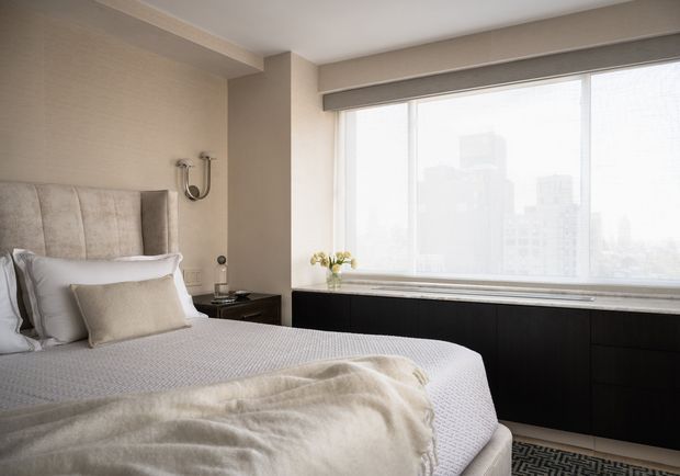 A bright, refined bedroom with a large upholstered bed, layered neutral bedding, and a textured throw. A dark built-in cabinet runs beneath expansive city-view glazing, complemented by a small bedside table and wall-mounted reading light. The palette is soft beige, cream, white, and charcoal, creating a calm contemporary hotel-like atmosphere.