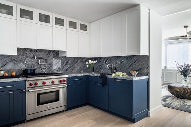 A bright, sophisticated kitchen with dark navy-blue lower cabinetry, white Shaker-style upper cabinets, and a dramatic grey veined stone backsplash and benchtop. The space includes a stainless-steel range cooker, undermount sink, integrated cabinetry, pale timber flooring, and a compact dining or living area visible beyond the opening.
