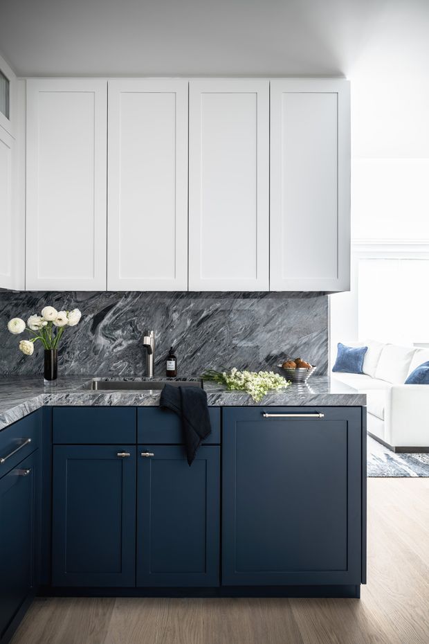 A refined kitchen interior featuring dark navy-blue shaker-style base cabinetry, white upper cabinets, and a dramatic grey-and-white veined stone backsplash and benchtop. A stainless-steel tap and undermount sink sit beneath the cabinetry, with simple brass-toned hardware. Styling includes flowers, a soap dispenser, fruit bowl, and folded dark textile. A bright white living area with blue cushions is visible beyond the kitchen.