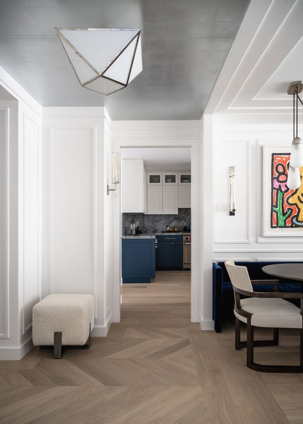 A refined transitional interior view from a paneled hallway into a compact kitchen and adjacent dining area. The space features white decorative wall panelling and trim, a grey plastered ceiling, herringbone timber flooring, dark navy kitchen cabinetry, a dark stone splashback, and a blue upholstered dining banquette with light upholstered chairs. Geometric pendant lighting and colourful framed artwork add visual interest.