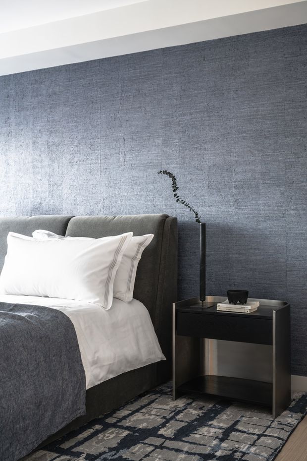 A refined bedroom vignette featuring a dark upholstered bed with crisp white bedding and a charcoal grey throw, set against a textured blue-grey feature wall. A black bedside table with a slim sculptural lamp sits beside the bed, while a patterned grey, black and cream rug grounds the room. The palette is cool, restrained and contemporary.