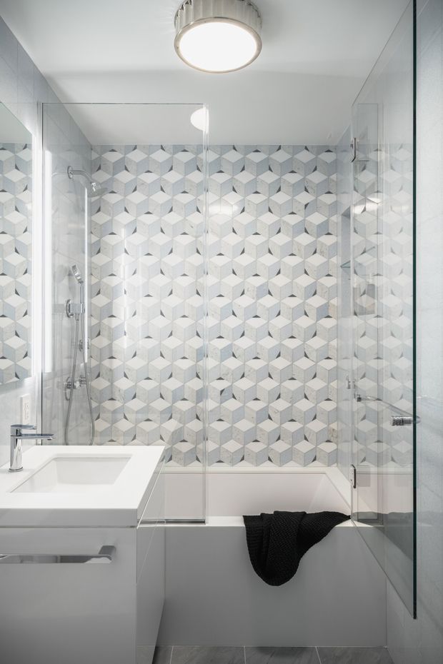 Bright contemporary bathroom featuring a white bathtub enclosed by clear glass shower screens, a white vanity with integrated basin, chrome tapware, and a striking geometric cube-pattern tiled feature wall in white and pale grey. The space has a clean, compact layout with a round ceiling light and grey floor tiles.