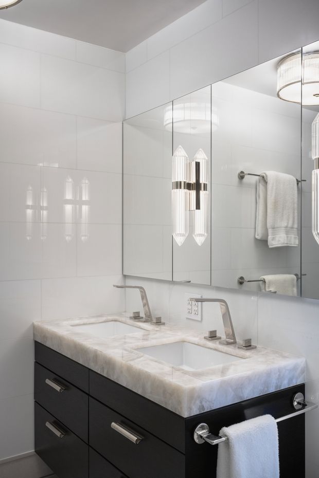 A bright, refined bathroom vanity with a double integrated sink, white marble-look benchtop, dark flat-panel cabinetry, polished chrome fixtures, large mirrored wall cabinets, white tiled walls, and sculptural wall sconces. The space has a clean, luxurious contemporary feel.