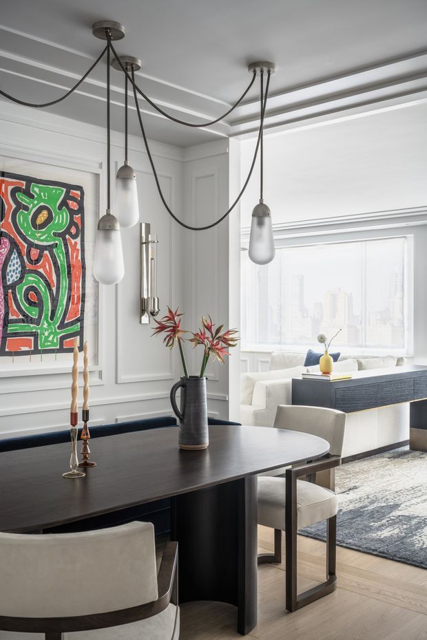 A refined open-plan dining and living area with a dark rectangular dining table, upholstered dining chairs, sculptural pendant lights with long black cords, and a decorative vase of branches. White walls feature detailed panel moulding, framed colourful artwork, and a large bright window. The adjacent living area includes a pale sofa, dark built-in cabinetry, a neutral rug, and light timber flooring. The overall composition is elegant, contemporary, and high-contrast.