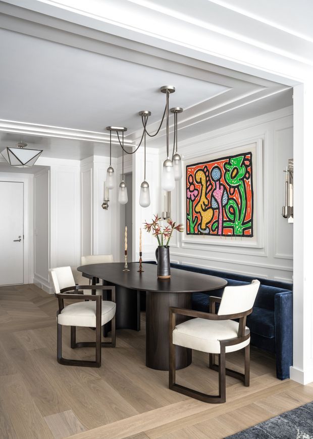 A refined dining area with a large dark oval dining table, upholstered wood-framed dining chairs, and a built-in deep navy banquette. The room features white decorative wall panelling, coffered ceiling detailing, layered pendant lighting, light timber flooring, and a colourful framed artwork. The overall composition blends classic architectural detailing with contemporary furniture and lighting.