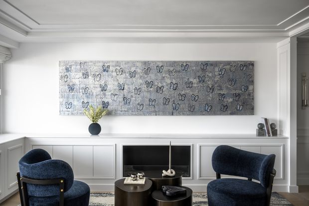Refined contemporary living room with white panelled built-in cabinetry, a recessed fireplace or television niche, dark blue upholstered armchairs, nesting coffee tables, and a large monochromatic abstract artwork. The room features a detailed stepped ceiling, white architectural trim, and a restrained navy, grey, and white palette.
