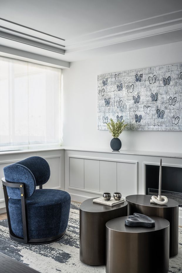 Refined contemporary living room with a bright white and grey palette, custom wall panelling, a built-in fireplace and television surround, layered recessed ceiling details, and a large butterfly artwork. A deep navy sculptural armchair contrasts with three dark cylindrical coffee tables, while sheer curtains diffuse abundant daylight.