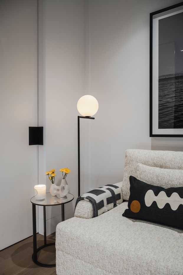 A close-up interior view of a softly styled living room corner, featuring a light textured upholstered sofa with graphic black-and-white cushions, a slim black metal side table with decorative vases and a candle, and a minimalist black floor lamp with a spherical globe shade. The room has pale grey walls, dark timber flooring, black-framed monochrome artwork, and a clean contemporary aesthetic.