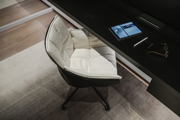 Close-up interior view of a refined home office or workspace, featuring a sculptural upholstered swivel chair beside a sleek black built-in desk. A tablet, stylus and glasses rest on the desktop, with neutral carpeting, timber flooring and dark contemporary joinery visible.