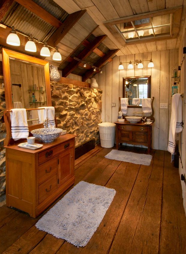 A rustic bathroom with extensive timber wall and ceiling panelling, wide-plank timber flooring, exposed dark timber beams, and a rough natural-stone feature wall. Two traditional timber vanities with vessel basins, ornate mirrors, white towels, pendant lighting, and a small skylight create a warm, vintage lodge-like atmosphere.
