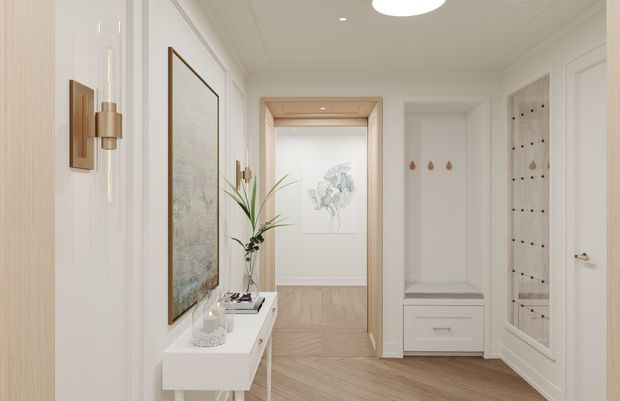 Bright, refined residential hallway or entryway with white paneled walls, decorative trim, light timber herringbone flooring, and a built-in console beneath a large framed artwork. The space includes a recessed built-in bench or storage unit and a glass-fronted display or wardrobe cabinet, with warm brass and timber accents, integrated lighting, and a view toward an adjoining room.