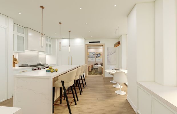Bright, elongated contemporary kitchen with white paneled cabinetry, a large white island with integrated seating, stainless-steel appliances, pendant lighting, and warm timber-look flooring. The open layout extends toward a small dining or secondary seating area, with built-in cabinetry along the right wall.