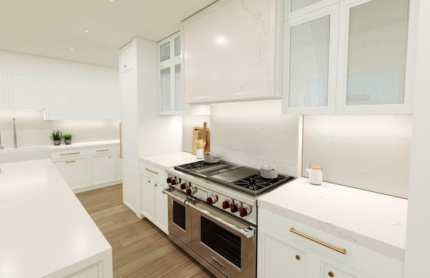 Bright, white traditional-style kitchen with extensive shaker-style cabinetry, white stone-look benchtops and splashback, brass-toned hardware, glass-front upper cabinets, and a large stainless-steel range cooker. Light timber-look flooring adds warmth to the otherwise crisp, airy space.