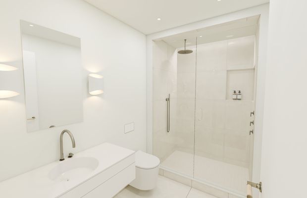 Bright, minimalist white bathroom with a floating vanity, integrated basin, wall mirror, wall-mounted lighting, toilet, and a frameless glass walk-in shower with overhead rain shower and recessed niches.