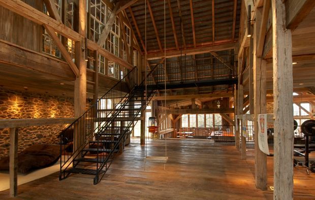 A large rustic barn-like interior with exposed timber posts, beams and rafters, weathered wood wall cladding, a broad timber floor, and a black metal open staircase leading to a mezzanine. A rough stone feature wall and extensive glazing add texture and daylight, while the open structural frame creates an industrial-rustic character.
