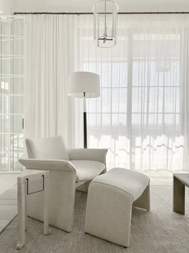 Bright, airy sitting area with a sculptural upholstered armchair and matching curved ottoman, positioned on a neutral textured rug. Sheer floor-to-ceiling curtains cover large gridded windows, while a white glass-front cabinet, slender floor lamp, contemporary pendant light, and simple side table complete the refined monochromatic interior.