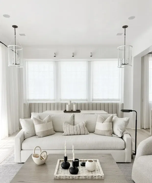 Bright, refined living room with a large cream upholstered sofa, layered neutral cushions, a matching curved banquette beneath three tall windows, sheer white curtains, and a pale wood coffee table styled with minimalist black and white ceramics. White walls and ceiling, slim black floor lamp, and sculptural pendant lights create an airy, contemporary setting.