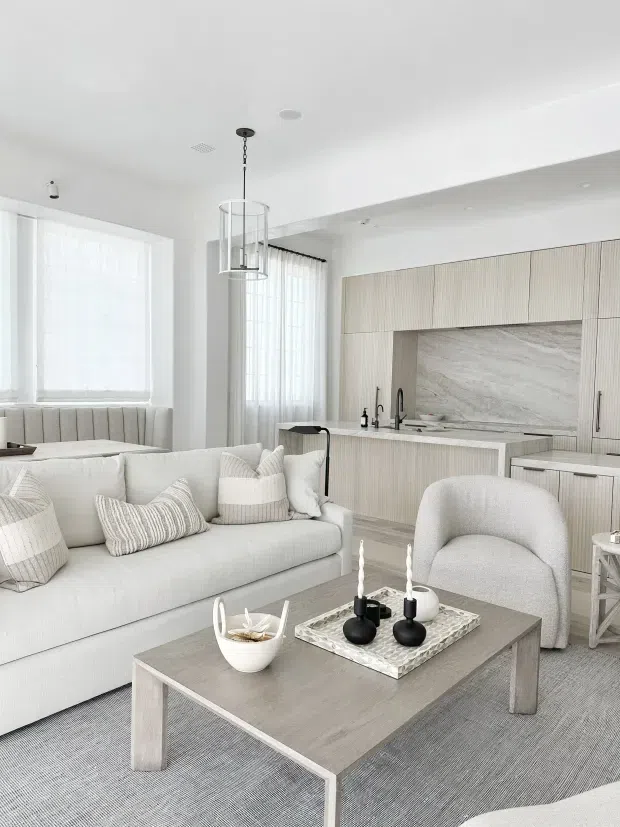 Bright open-plan living room and kitchen with a soft neutral palette. The room features a large light upholstered sofa, textured cushions, sculptural armchair, pale timber coffee table, grey carpet, built-in dining nook, and a contemporary kitchen with vertically fluted light cabinetry, integrated island, dark tapware, and a large natural stone-look splashback. Sheer curtains and restrained black accents create an airy, refined interior.