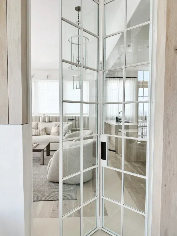 Bright contemporary interior viewed through a white-framed, multi-pane glazed pivot or folding door. The foreground opens between a light-toned hallway or entry area and an airy living room, with pale upholstered seating, a low dark timber coffee table, and a textured rug. A kitchen with a stone island and dark tapware is visible beyond the living space. Light timber wall cladding and flooring, white walls, sheer curtains, and pendant lights create a refined, cohesive open-plan interior.