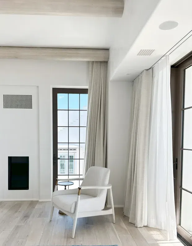 Bright, refined sitting area with a white upholstered lounge chair positioned beside expansive black-framed glazed doors and windows. Full-height sheer and linen curtains soften the light, while pale timber ceiling beams and matching light timber flooring create a warm, understated interior. A wall-mounted television and discreet linear ventilation grilles are visible.