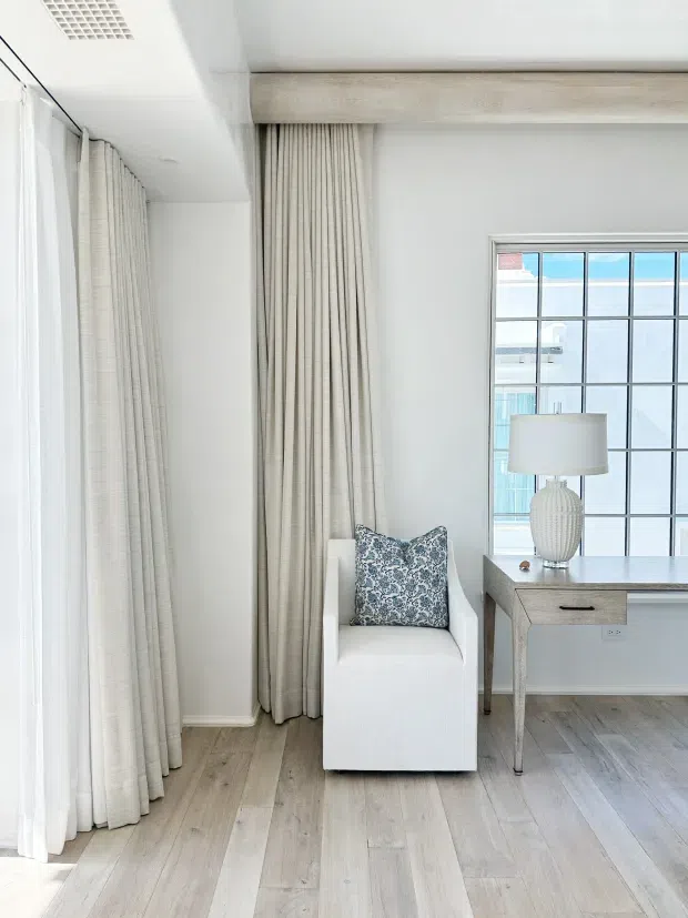 Bright, refined interior corner featuring a white upholstered armchair with a patterned blue-and-white cushion, floor-to-ceiling neutral curtains, pale timber flooring, and a light timber desk beneath a large black-grid window. A textured ceramic lamp sits on the desk, with a coastal exterior visible through the glazing.