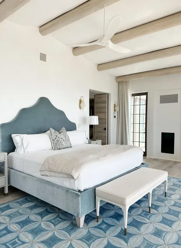 Bright, refined bedroom with a large upholstered bed featuring a sculptural blue-grey headboard, white bedding, and a light upholstered bench. The room has a patterned blue and beige carpet, white walls, pale timber ceiling beams, a white ceiling fan, decorative wall sconces, full-height curtains, and a glazed exterior door. A dark fireplace opening is visible on the right wall.