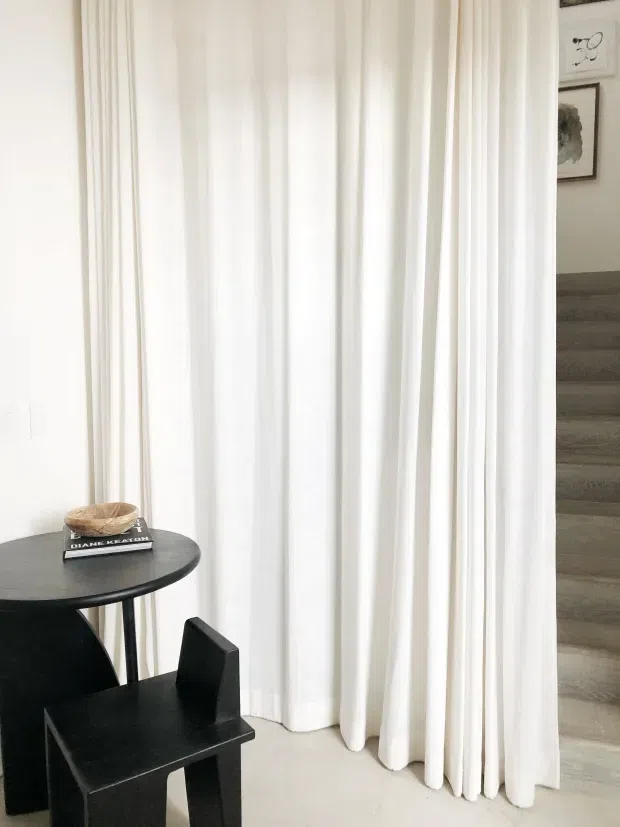 A bright, minimalist interior corner with full-height white sheer curtains, a small round black side table, and a sculptural black chair. A grey-toned timber staircase rises at the right, with framed artwork visible on the adjacent wall. The space has a clean, restrained palette and strong vertical drapery lines.