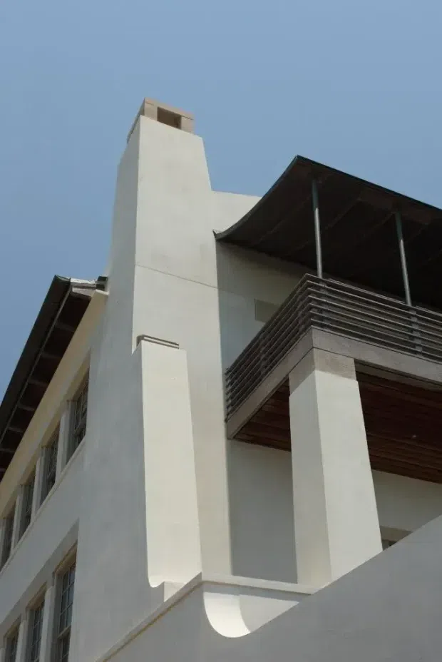 Low-angle exterior photograph of a multi-level modern building with white rendered walls, strong vertical chimney-like projections, deep roof overhangs, slender metal supports, timber soffits, and a dark horizontal slatted balcony balustrade. Clear blue sky forms the background.
