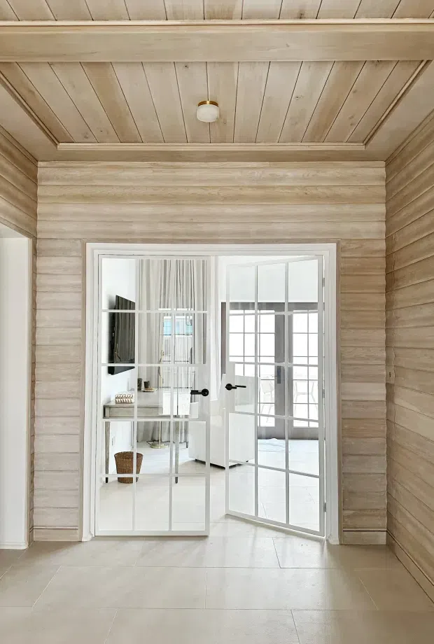 Bright transitional interior view focused on a white-framed, glazed French-style double door with black hardware. The surrounding walls and ceiling are clad in pale natural timber boards, with a recessed timber ceiling panel and exposed trim. Pale large-format floor tiles continue through the foreground, while a light, airy room is visible beyond the open doors.