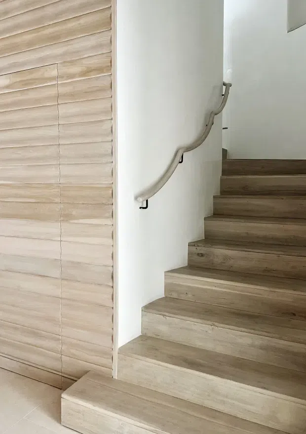 Interior view of a modern stairway with broad pale timber-finished treads and risers, a smooth white wall, and a curved light-coloured handrail with dark brackets. The adjacent wall is clad in horizontal light timber boards, creating a warm, minimal staircase composition.